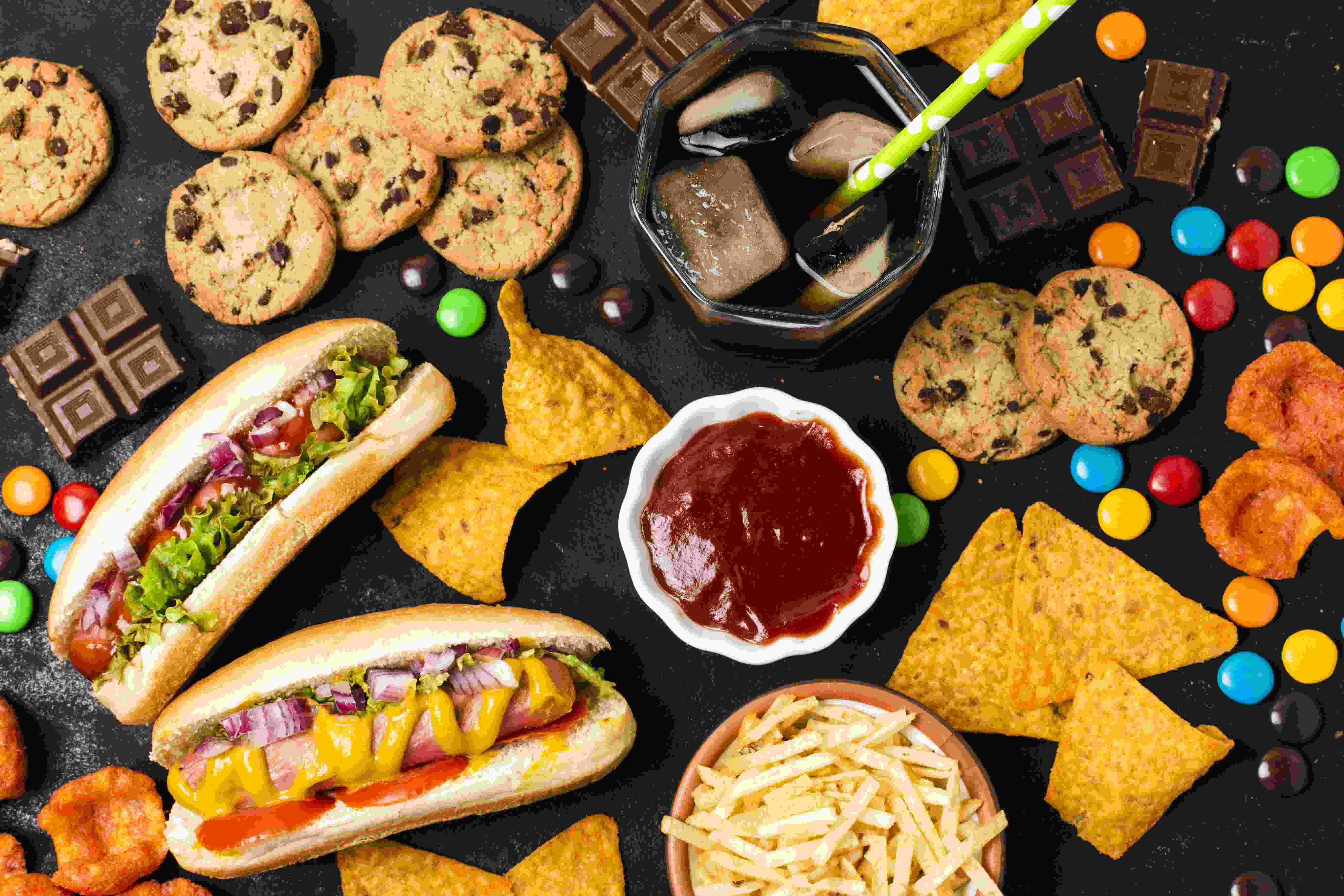 Ultra-processed food including hot dogs, cookies, chips, and soda on a dark background.